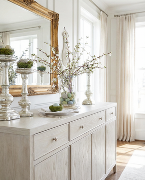 Light wood dresser with decorative items including a large mirror, vases, and branches.